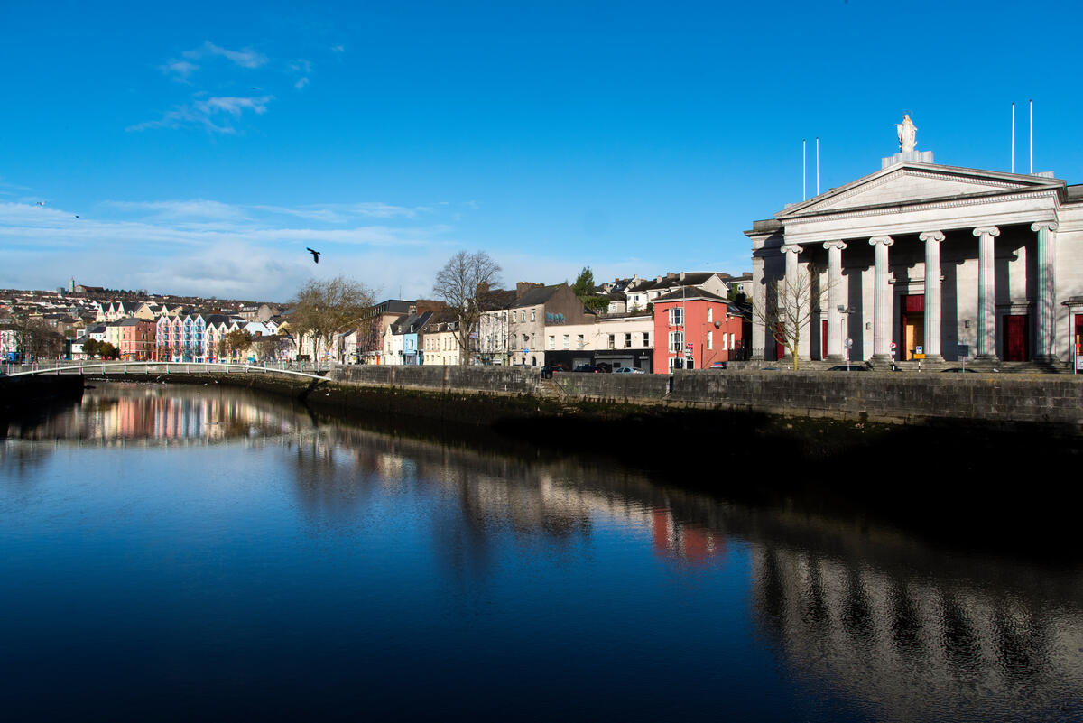The River Lee reflecting Cork city buildings and St Mary&rsquo;s Priory on Pope&rsquo;s Quay on a clear blue day.